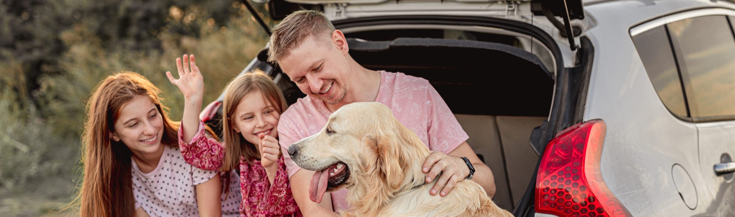 A father with his daughters and dog sitting in an open car trunk outdoors.