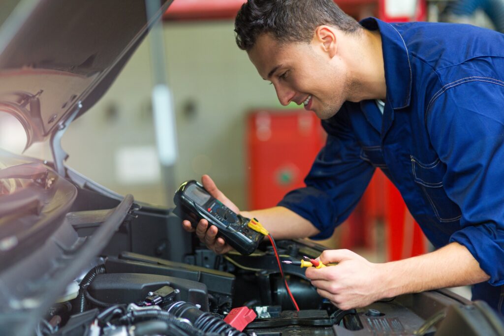 A mechanic in a blue uniform tests a car battery using a diagnostic tool, ensuring the vehicle is ready for springtime driving.