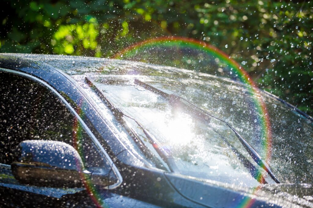 Water sprays off a car windshield, creating a rainbow effect in the sunlight, symbolizing the freshness of spring car maintenance.