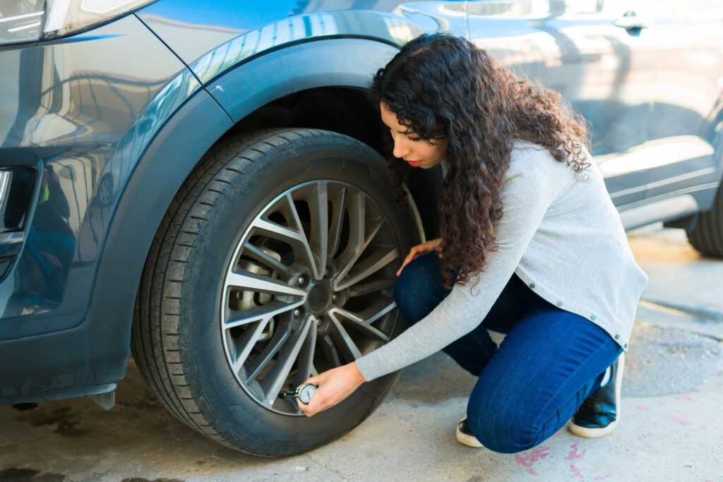 A woman crouches beside her car, checking the tire pressure with a gauge to ensure safe spring driving and proper vehicle maintenance.