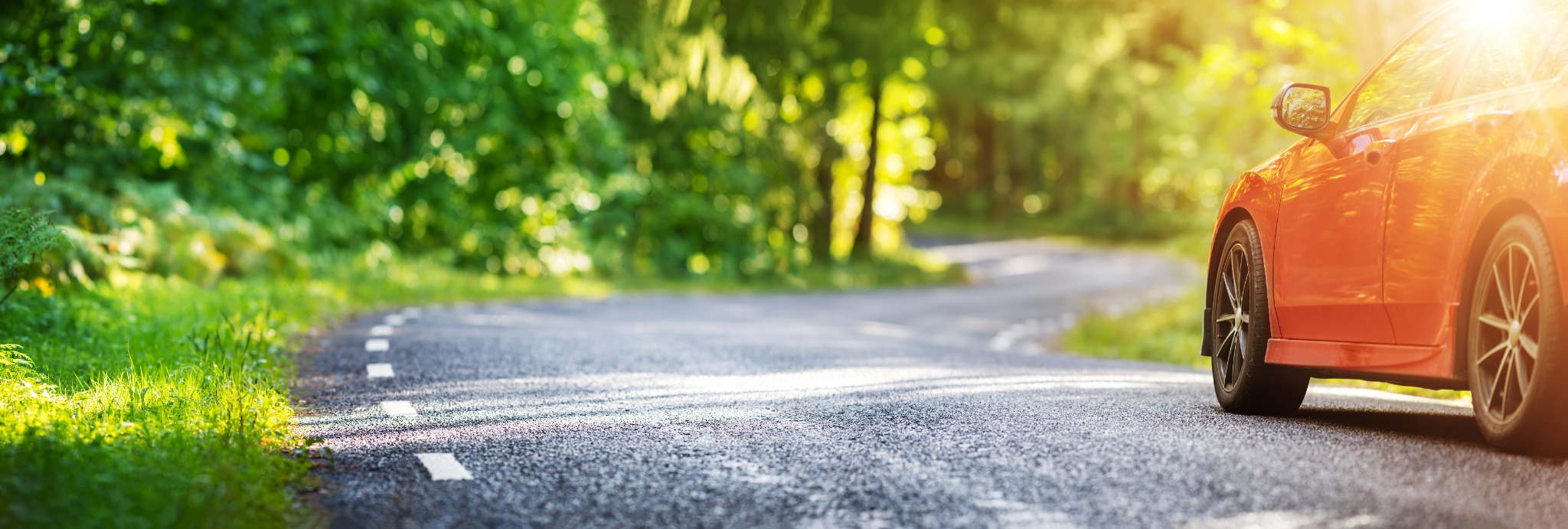 A red car drives down a winding asphalt road surrounded by lush greenery and sunlight, symbolizing spring car maintenance and seasonal driving tips.