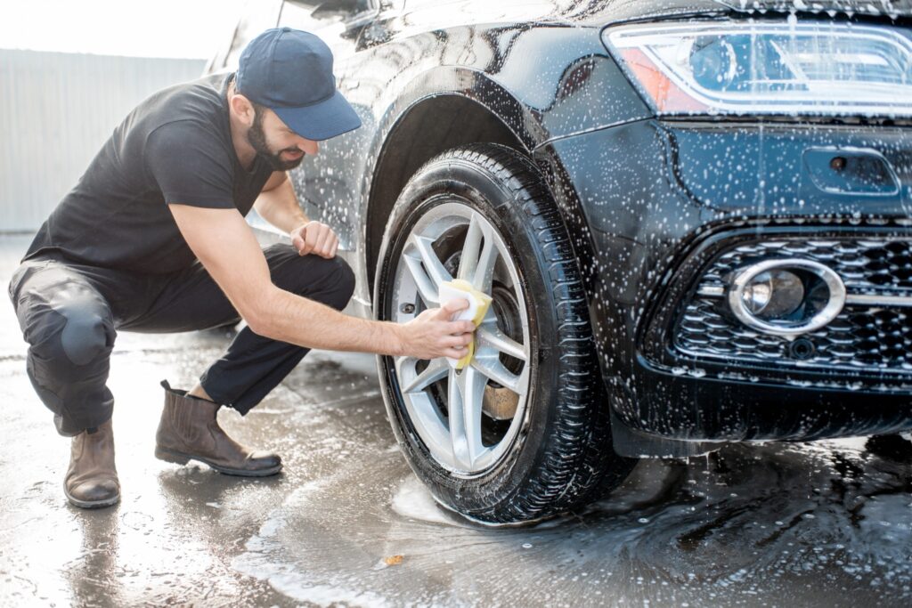 A man washes the tires of a black car, scrubbing off dirt and road salt as part of a spring vehicle cleaning routine.