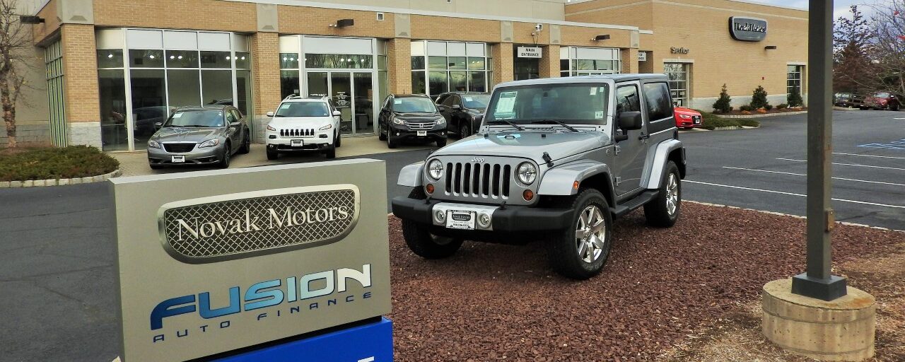Front view of Novak Motors dealership in Lebanon, New Jersey, with a silver Jeep parked beside a Fusion Auto Finance sign, marking the 2015 expansion of the Lease Return & Resale Center.